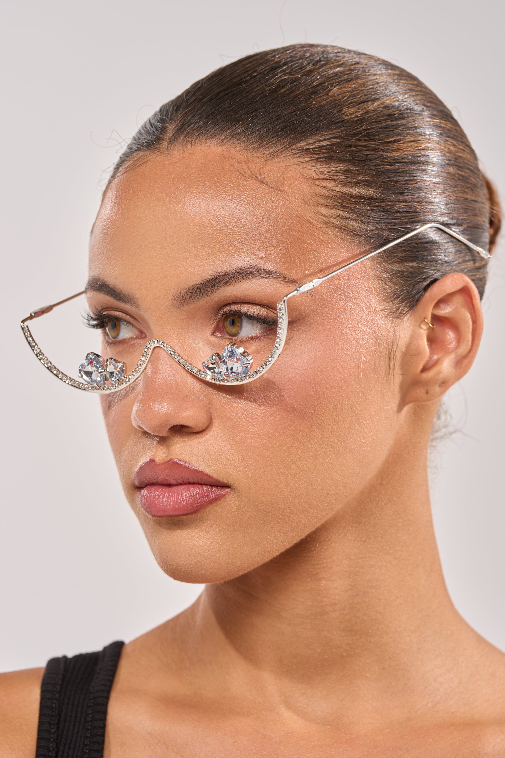 A woman with smooth, pulled-back hair wears DIAMOND DRIFT SUNGLASSES adorned with large, clear rhinestones beneath each eye. She has a neutral expression and is dressed in a black top. 0