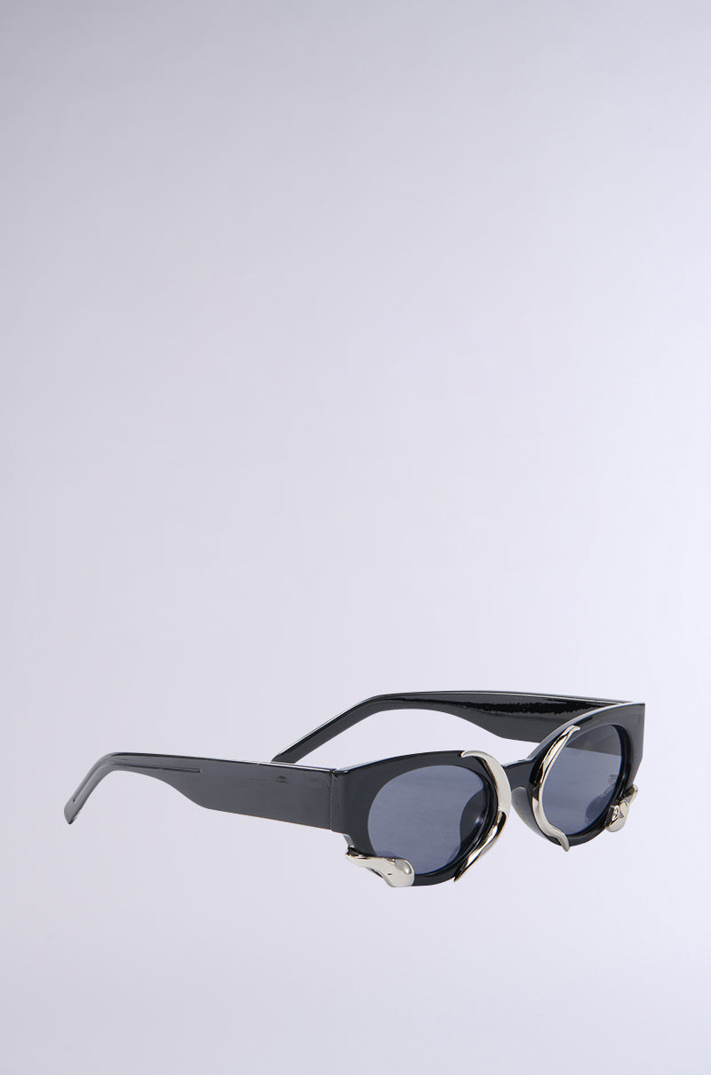 The VENOM LOVE SUNGLASSES are stylish black cat eye frames featuring dark lenses and unique metallic snake-inspired detailing, angled to showcase both the side and front views against a gray background.