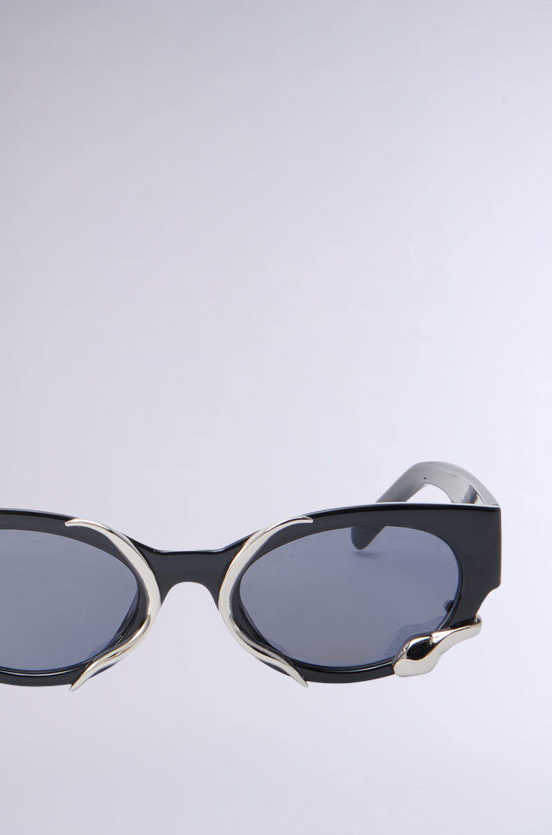 The VENOM LOVE SUNGLASSES, featuring sleek black cat-eye frames with silver side and top accents and metallic snake-inspired detailing on a light gray background, boast dark tinted lenses.