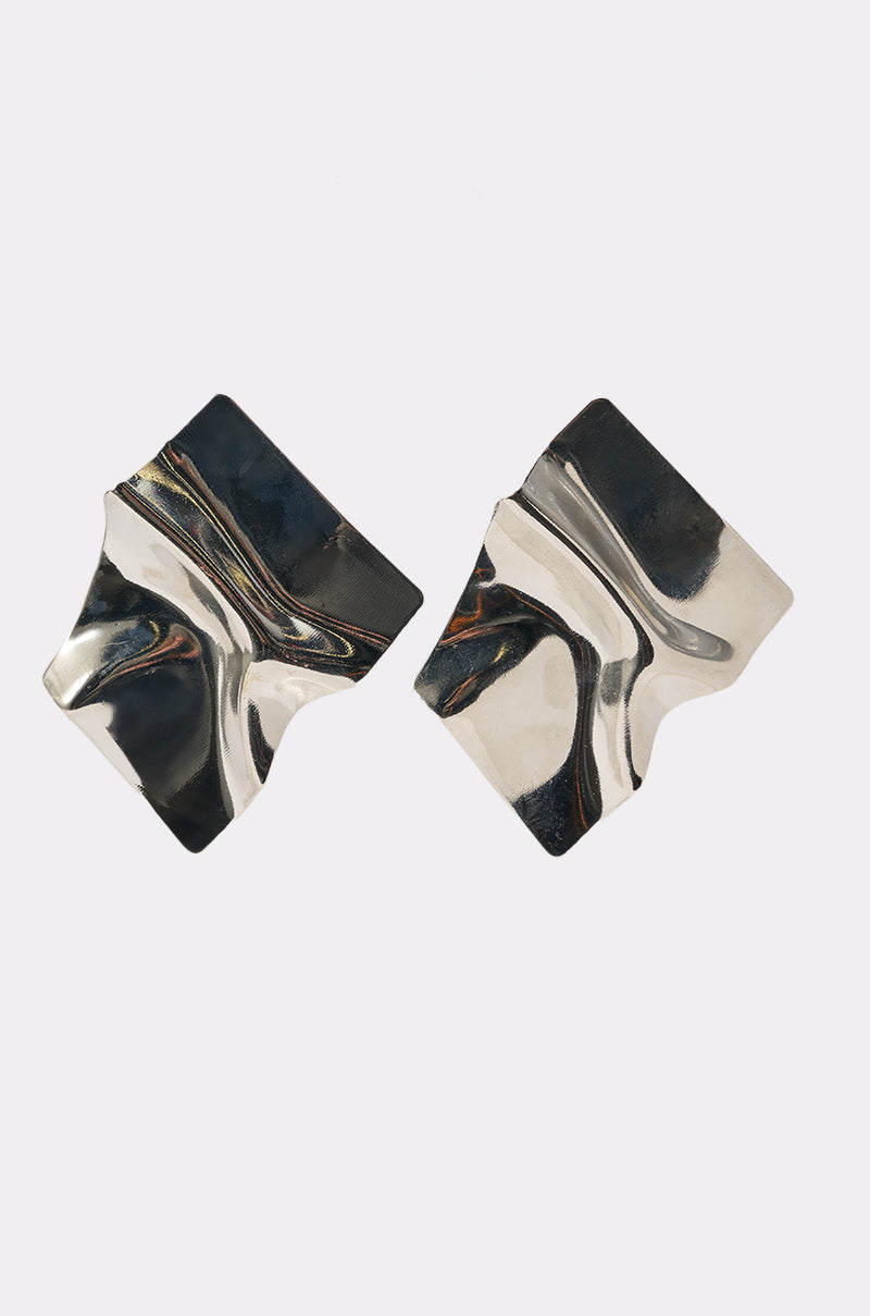 The MIRROR MIRROR OVERSIZED STUDS are faux silver, asymmetrical earrings with a wavy, folded design shown on a plain white background. 1