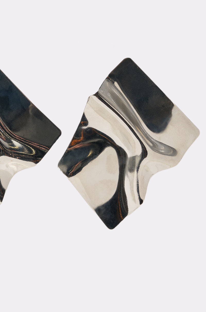 Two irregular, shiny metallic shapes with wavy surfaces are shown on a plain white background. The abstract forms are the MIRROR MIRROR OVERSIZED STUDS, featuring a fluid, reflective design and mirror-like finish. 3