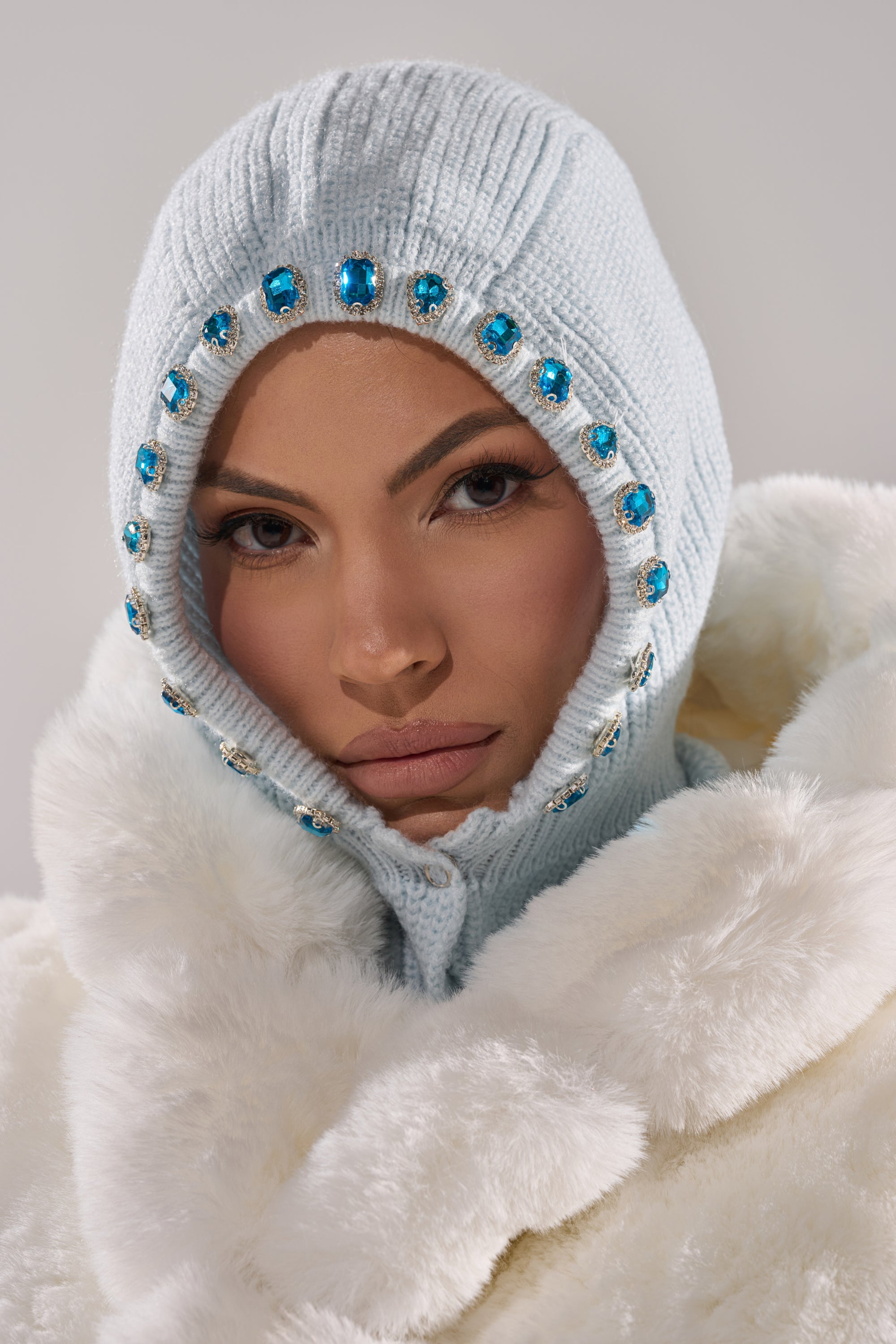 Wearing the TRU EMBELLISHED SNOOD—a light blue fisherman knit hood with rhinestone jewels—and a white faux fur coat, a person looks directly at the camera with a neutral expression. 1