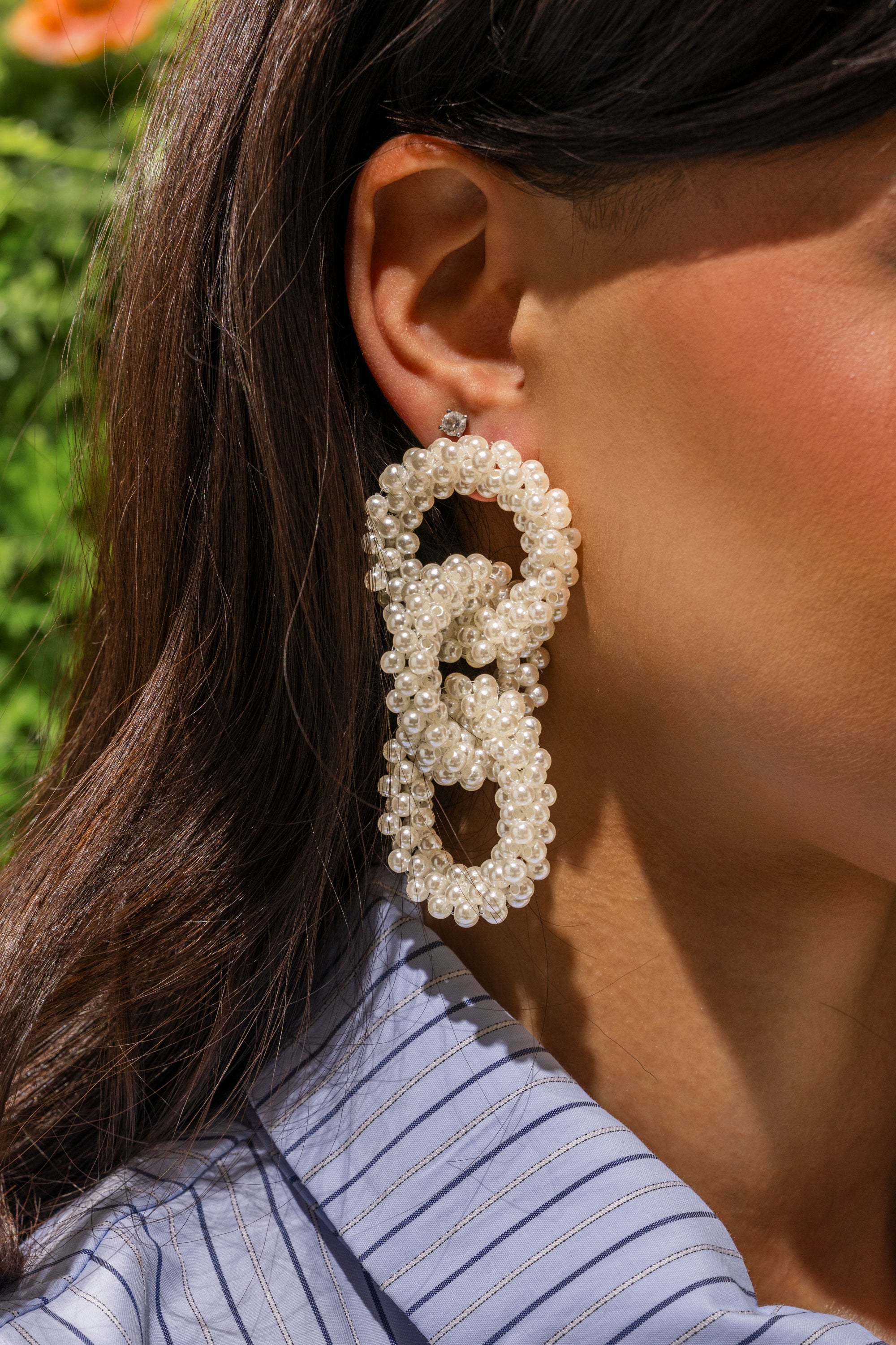 A close-up shows a woman with long brown hair in a blue pinstriped shirt wearing PEARL BABY DANGLE EARRING, featuring interlinked loops with small pearls. Greenery appears in the background. 0