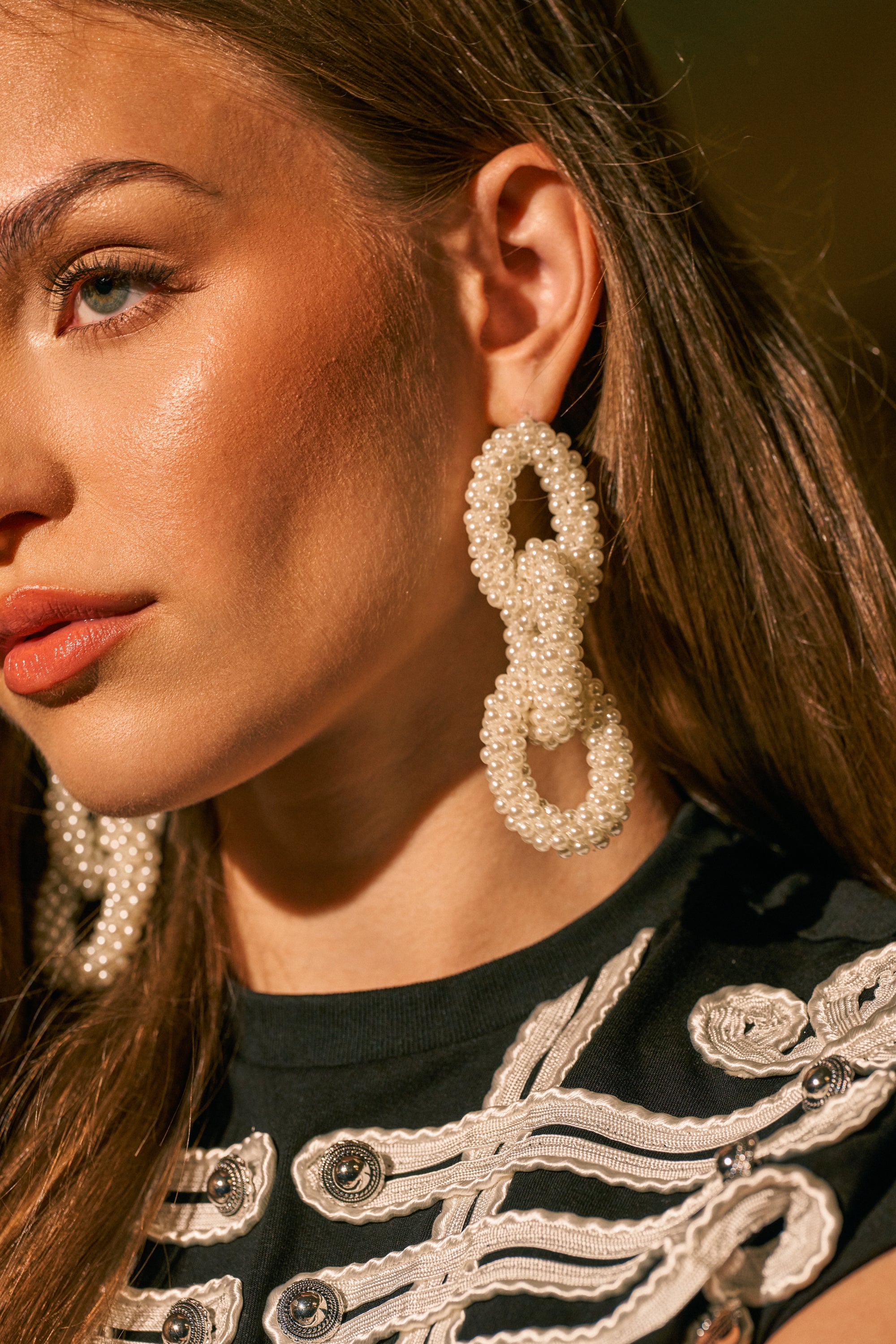 A woman with straight brown hair models the PEARL BABY DANGLE EARRING—large, oval-shaped pearl drop earrings—paired with a black top featuring white decorative embroidery, all shown in warm lighting.