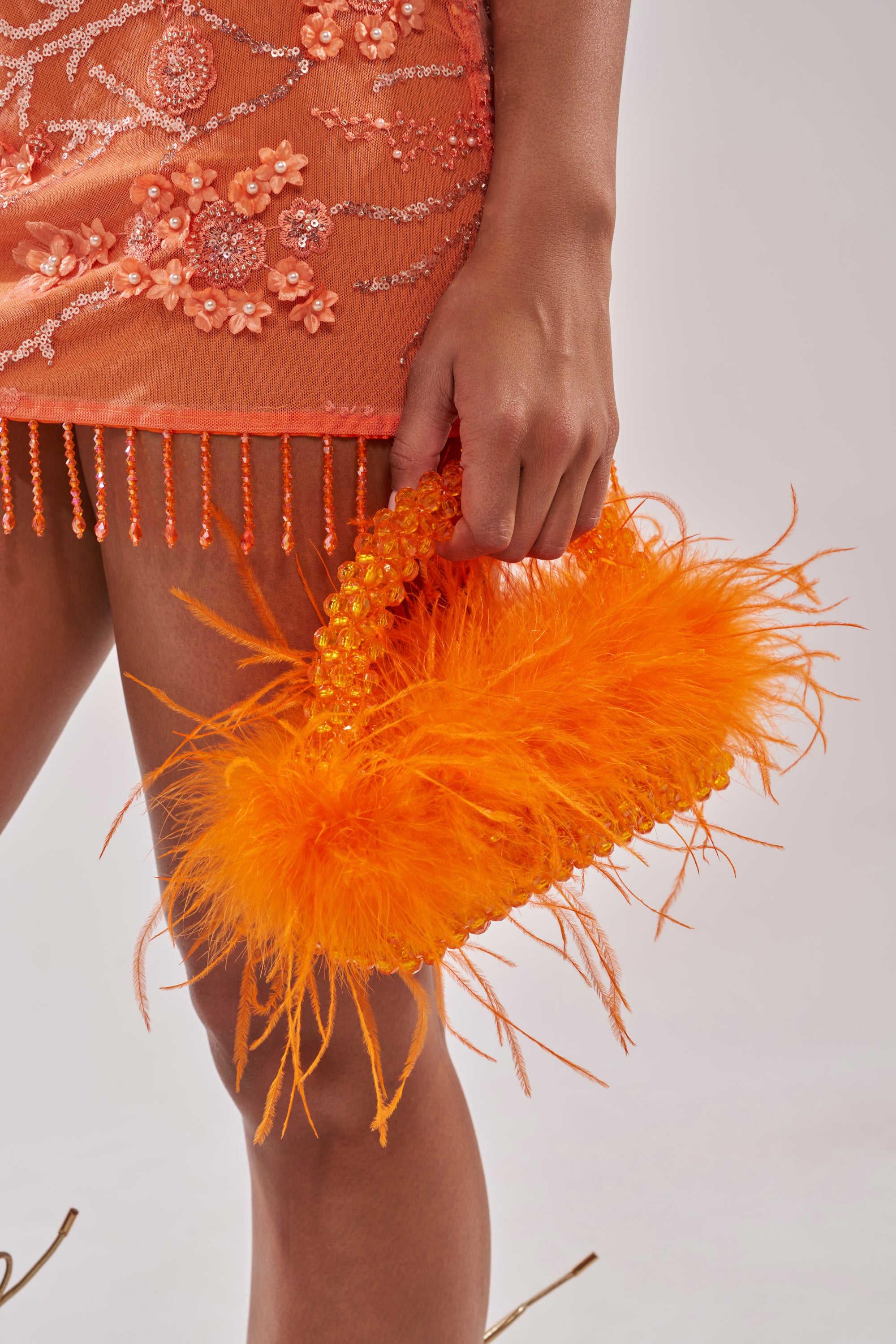 A person in an embellished orange dress holds the NIGHT OUT BEADED FEATHER BAG, a vibrant orange handbag adorned with delicate beads and fluffy feathers. 1
