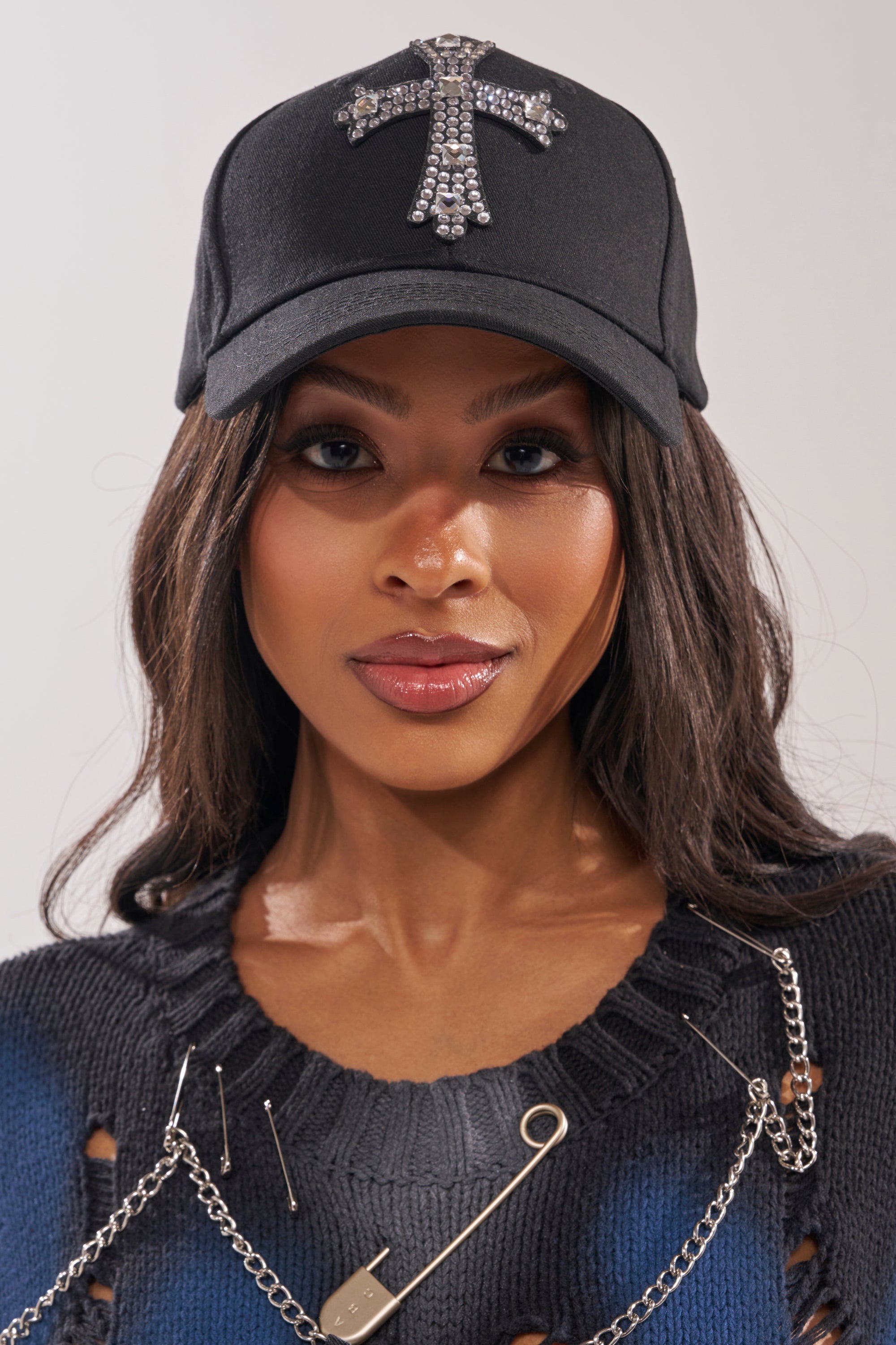 A woman in the EMBELLISH ME BASEBALL HAT, paired with a distressed black sweater featuring safety pins and a chain, looks directly at the camera. 0