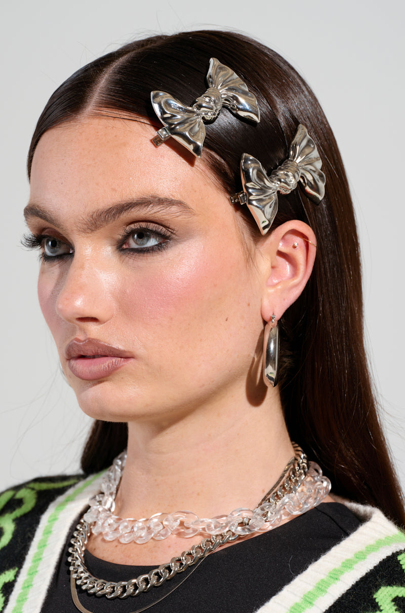 A person with long dark hair accessorizes with Bow Bow Metal Hair Clips, adding a touch of glamour to their look. Their bold eye makeup and light lipstick complement layered chain necklaces and large hoop earrings, while an intricately patterned garment teases from the shoulder.