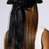 A person with long, straight hair wears the ROSA LACE BOW SNAPBACK, a black satin cap adorned with a large lace bow applique and ribbons cascading from the back. They are dressed in a sleeveless white top and are facing away from the camera, showcasing the bow and their hair.