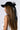 A person with long, straight brown hair seen from the side is sporting a ROSA LACE BOW SNAPBACK in black satin, featuring a large decorative lace bow applique and an adjustable strap at the back. They are dressed in a sleeveless white top against a plain white background.