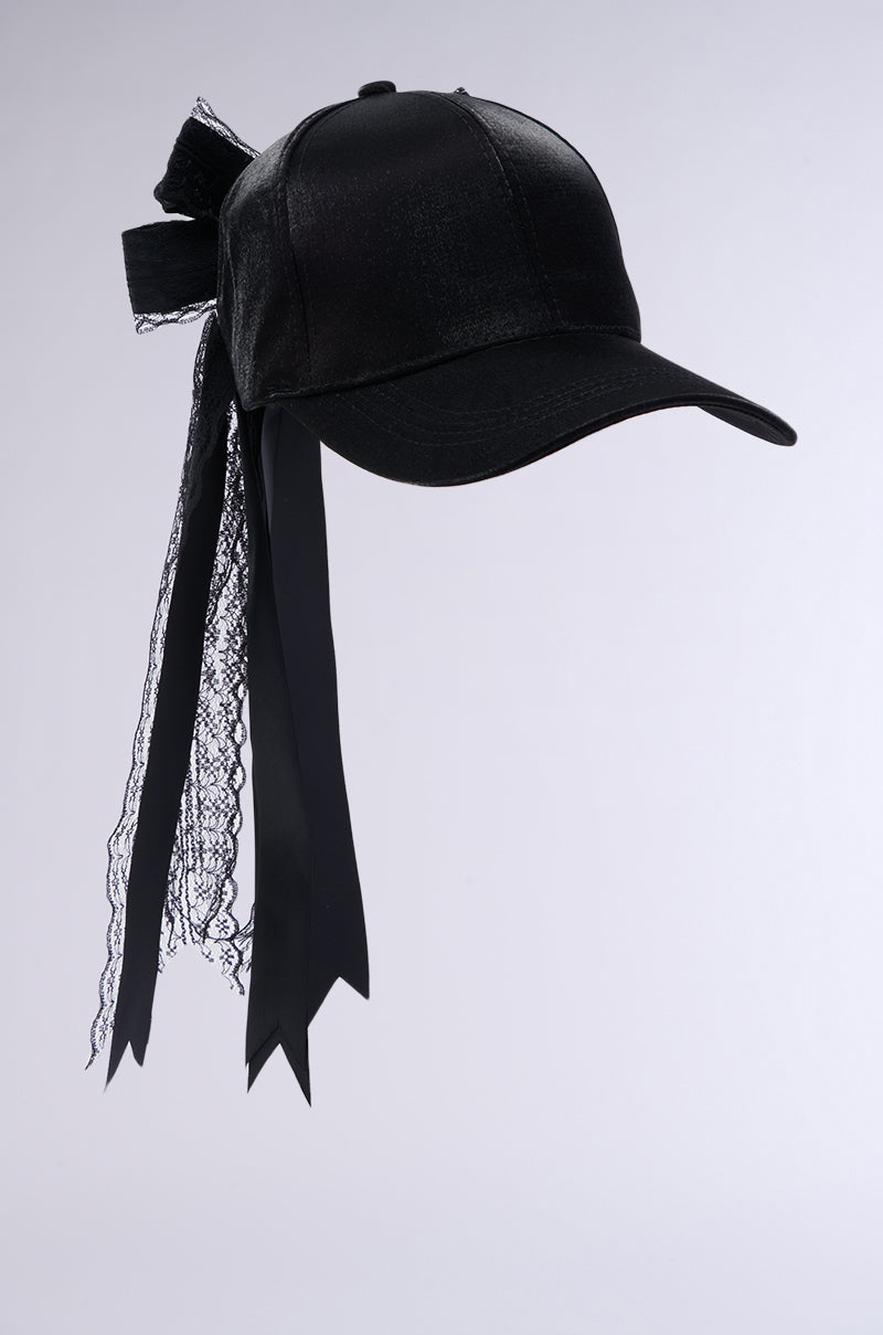 The ROSA LACE BOW SNAPBACK is a black baseball cap adorned with a large black bow and ribbon streamers cascading from the back. The streamers, made of a blend of shiny material and lace, provide a decorative and elegant touch to the otherwise casual cap. An adjustable strap ensures the perfect fit. The background is plain and light gray. 3