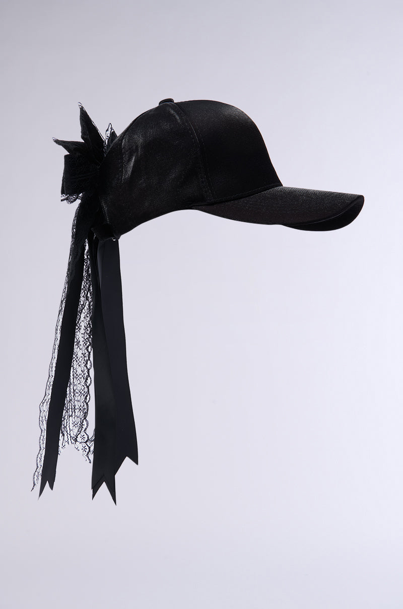 The ROSA LACE BOW SNAPBACK features a velvety black baseball cap adorned with a charming lace bow applique and flowing satin ribbons down the back. With an adjustable strap for a perfect fit, it boasts a clean, modern design set against a sleek gray background. 4
