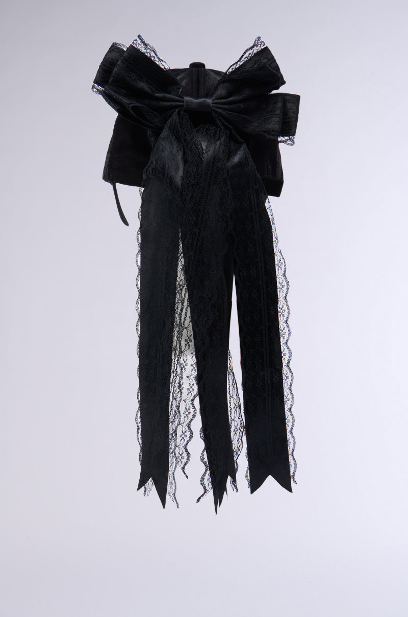 The ROSA LACE BOW SNAPBACK is a stylish black velvet bow embellished with intricate lace applique detailing. It boasts long, trailing ribbons edged with scalloped lace trim, making it an elegant and sophisticated accessory against a plain light background. 5