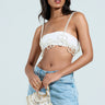 A person with long hair poses against a plain background, wearing a white embellished crop top with tank straps and light blue jeans, accessorized by the FLOWER BABE PEARL BODY CHAIN, which adds a touch of elegance to the ensemble.