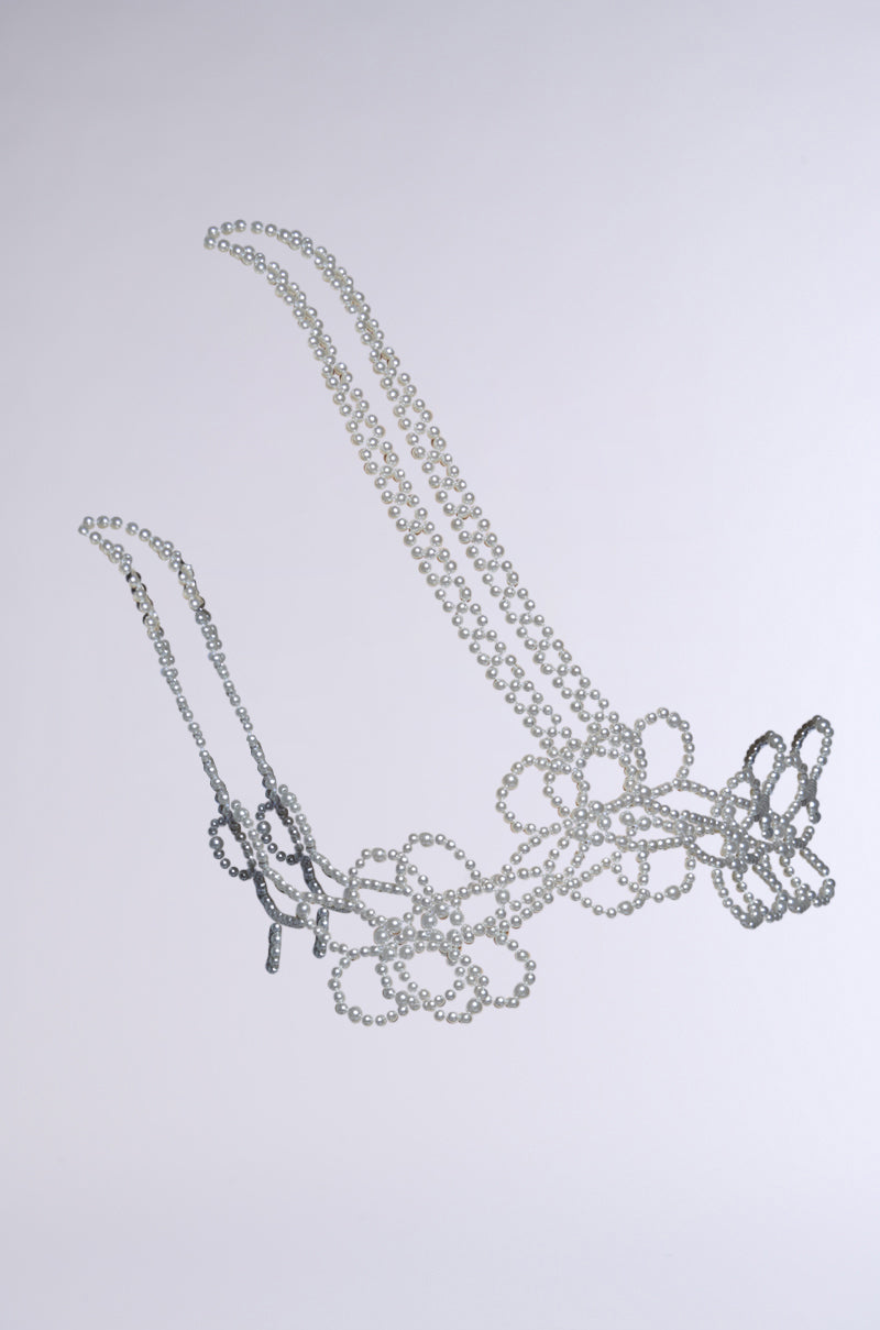 The FLOWER BABE PEARL BODY CHAIN features two parallel strands of interconnected silver loops shaped into floral patterns, each end adorned with floral pearl embellishments, elegantly draped against a light gray background.