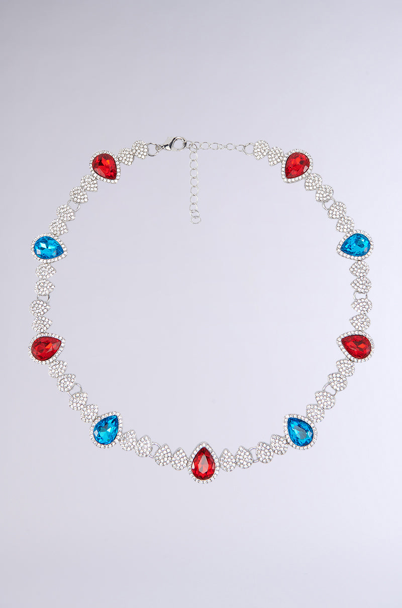 The TOO TURNT NECKLACE is a stunning faux silver accessory that showcases alternating red and blue teardrop-shaped gemstones, each accented with rhinestone embellishments and linked together by an intricate chain design. It features an adjustable clasp at the top for a perfect fit.