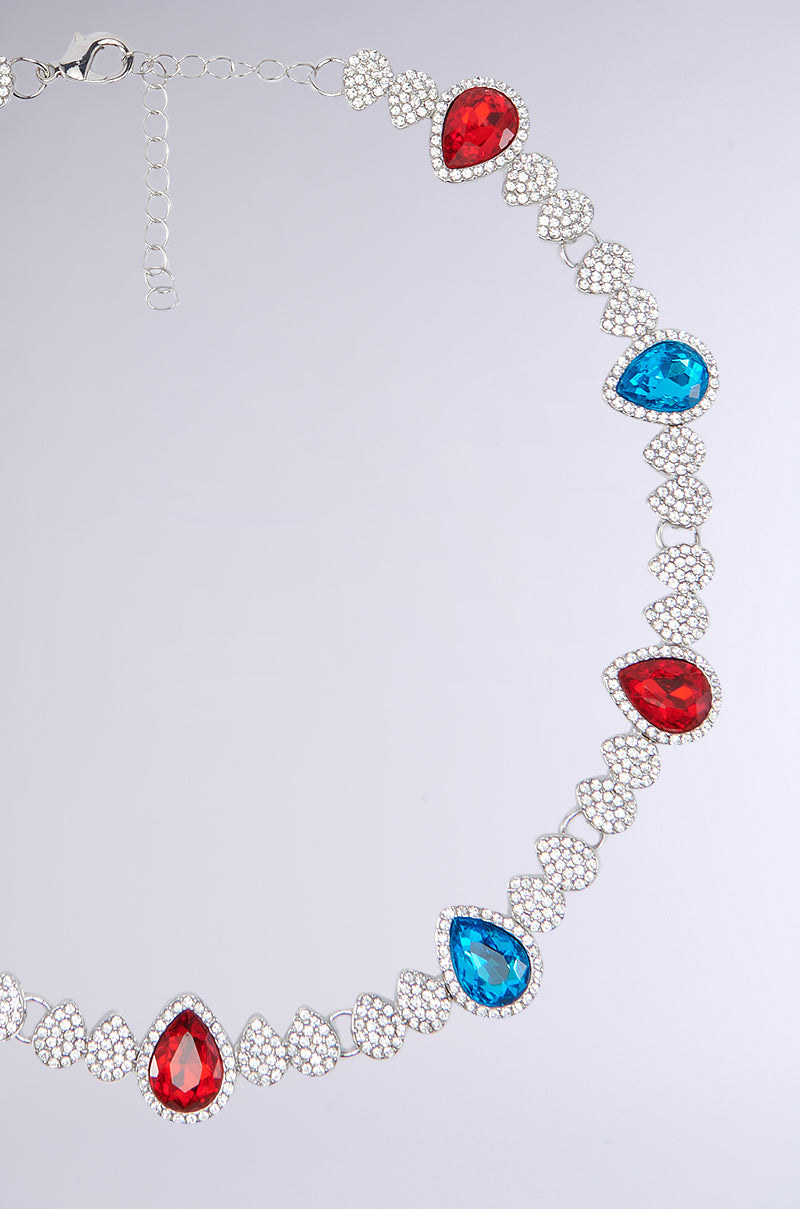 Introducing the TOO TURNT NECKLACE: a faux silver accessory featuring alternating red and blue teardrop-shaped gemstones, each meticulously set within a pear-shaped cluster of small clear crystals. This colorful piece is connected by a chain adorned with intricate heart-shaped designs and embellished with rhinestones, creating an elegant and vibrant statement.