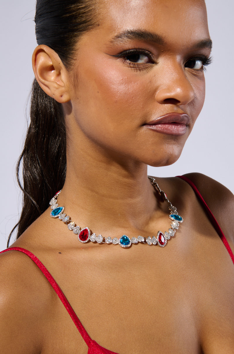 A woman wearing a red top and the TOO TURNT NECKLACE, featuring alternating blue and red gemstones separated by sparkling rhinestone embellishments. She has straight hair pulled back and is posing against a plain background, looking confidently at the camera.
