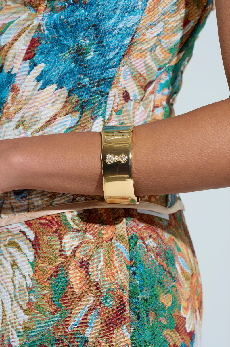 A person in a textured, colorful floral outfit is shown from the waist up, wearing the KEY TO MY HEART CUFF bracelet featuring a small ornate design. The background is light and plain.