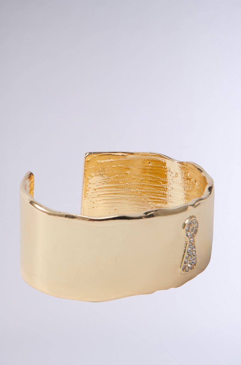 The KEY TO MY HEART CUFF is a metallic faux gold bracelet with a textured inner surface. Its outer side features a rhinestone-accented keyhole design with tiny, clear gemstones, creating an elegant minimalistic statement piece that stands out against neutral backgrounds.