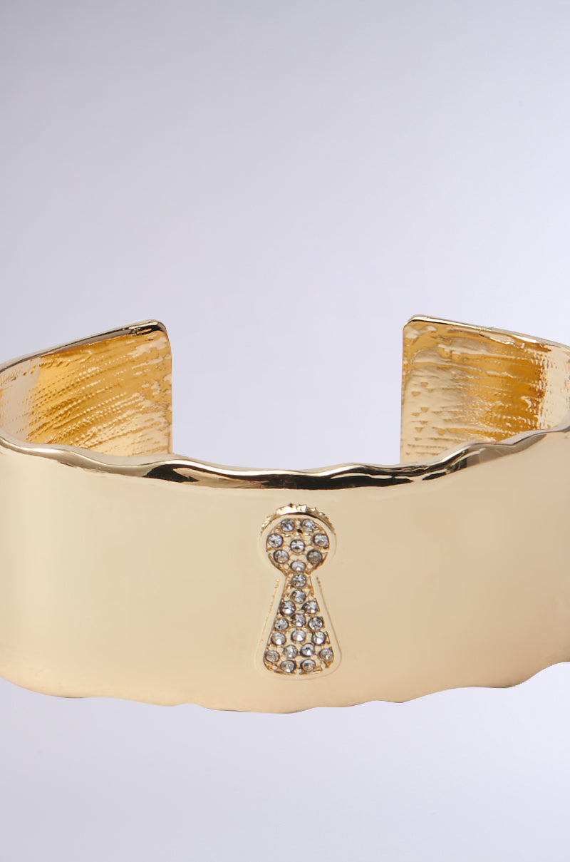 The KEY TO MY HEART CUFF is a chunky statement bracelet in metallic faux gold with a textured finish, featuring a rhinestone-adorned keyhole design at its center and an open back style for a luxurious and elegant appearance.