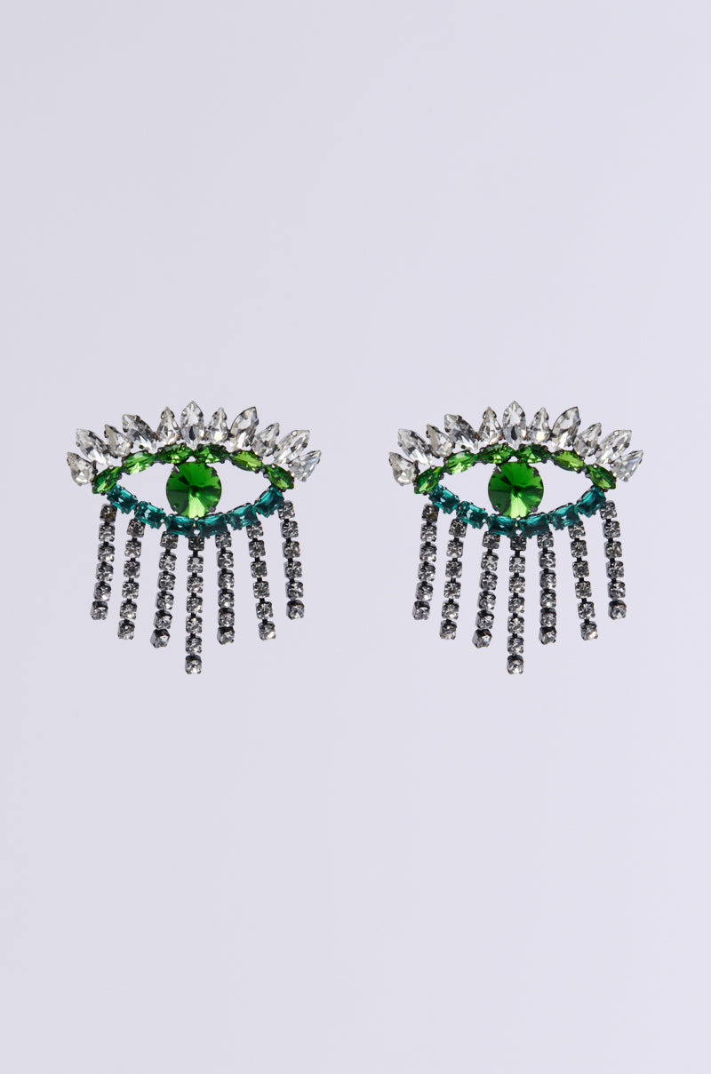 Close-up image of the EYE SEE YOU EARRING pair, showcasing a central green gemstone. They are elegantly framed by clear crystals and green accents, featuring rhinestone link fringes that mimic eyelashes. Set against a plain white background, these earrings are undeniably captivating.