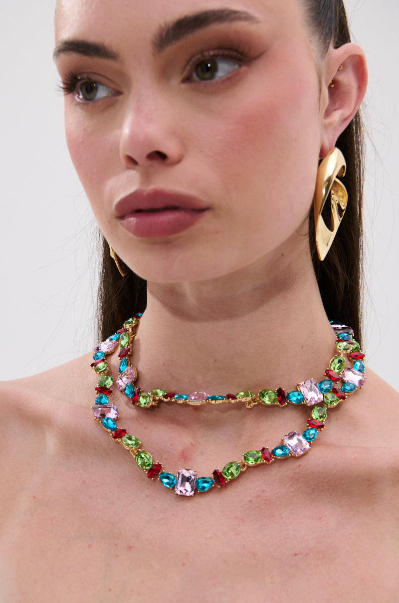A woman with fair skin and glossy lips wears the I WANT IT ALL NECKLACE, a bold piece featuring multicolored gemstones in pink, blue, and green, along with gold leaf-shaped earrings. She gazes slightly to the side.