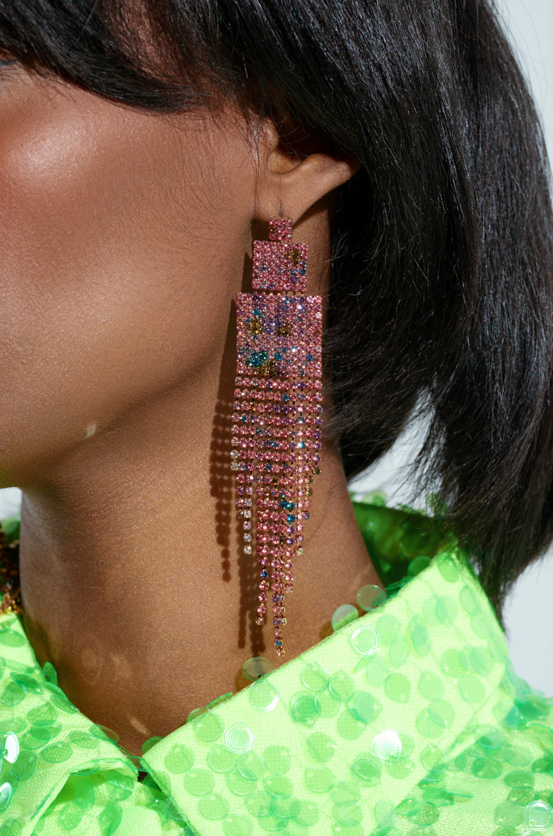 In a close-up shot, an individual showcases the stunning RAINBOW DREAMING DANGLE EARRING, adorned with multicolor rhinestone embellishments. The earring's shimmering, cascading design beautifully complements their vibrant green sequined outfit, adding an extra layer of glamour and sophistication.