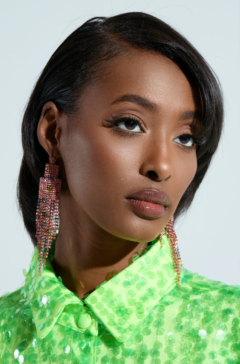 A person with short black hair wears a bright green sequined top and RAINBOW DREAMING DANGLE EARRINGS, gazing to the side. Their makeup is polished with bold eyelashes and glossy lips, set against a plain background, creating an enchanting yet sophisticated look.