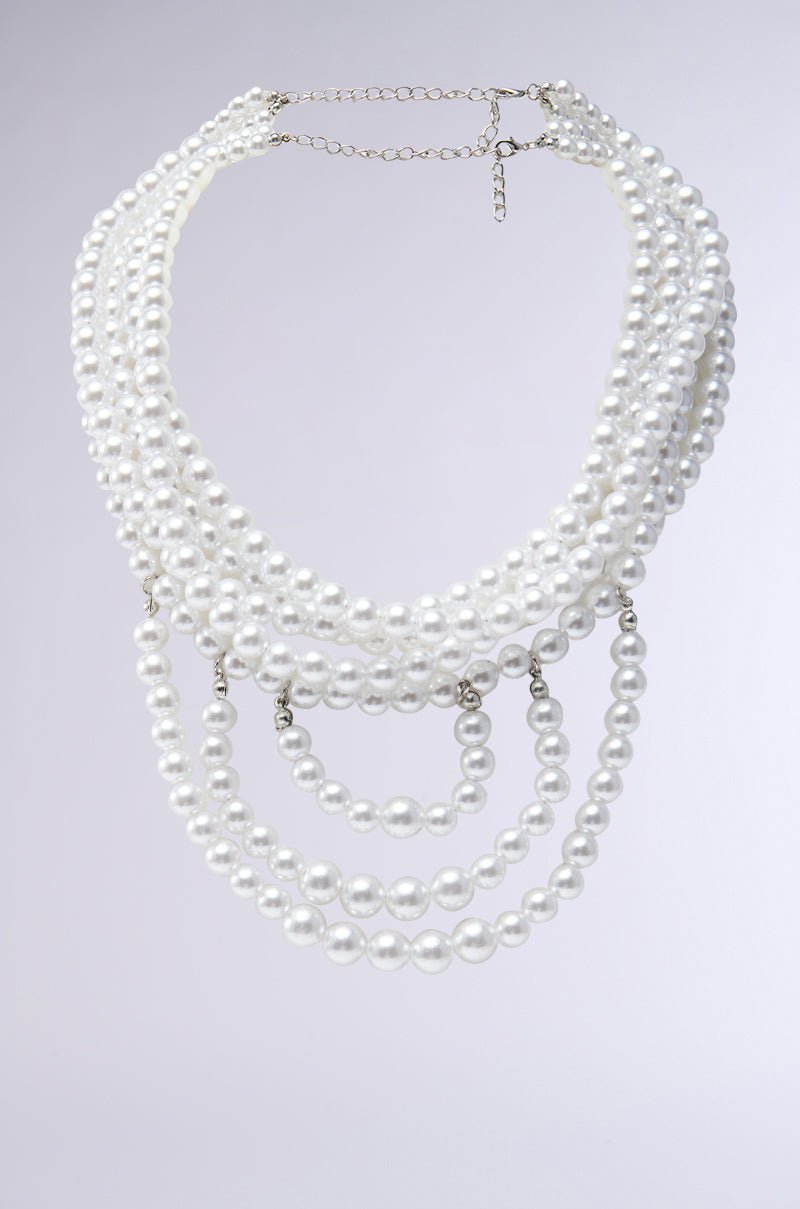 The ANNE LAYERED PEARL NECKLACE is a multi-strand piece with graduated white pearls set against a plain gray background, resembling a choker. The varied pearl sizes create a cascading look, attached to a fine chain with an adjustable clasp.
