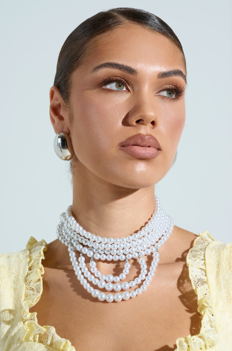 A woman with a sleek bun wears the ANNE LAYERED PEARL NECKLACE, silver hoop earrings, and a light yellow lace top. She looks to the side against a light background, exuding a polished and elegant appearance.