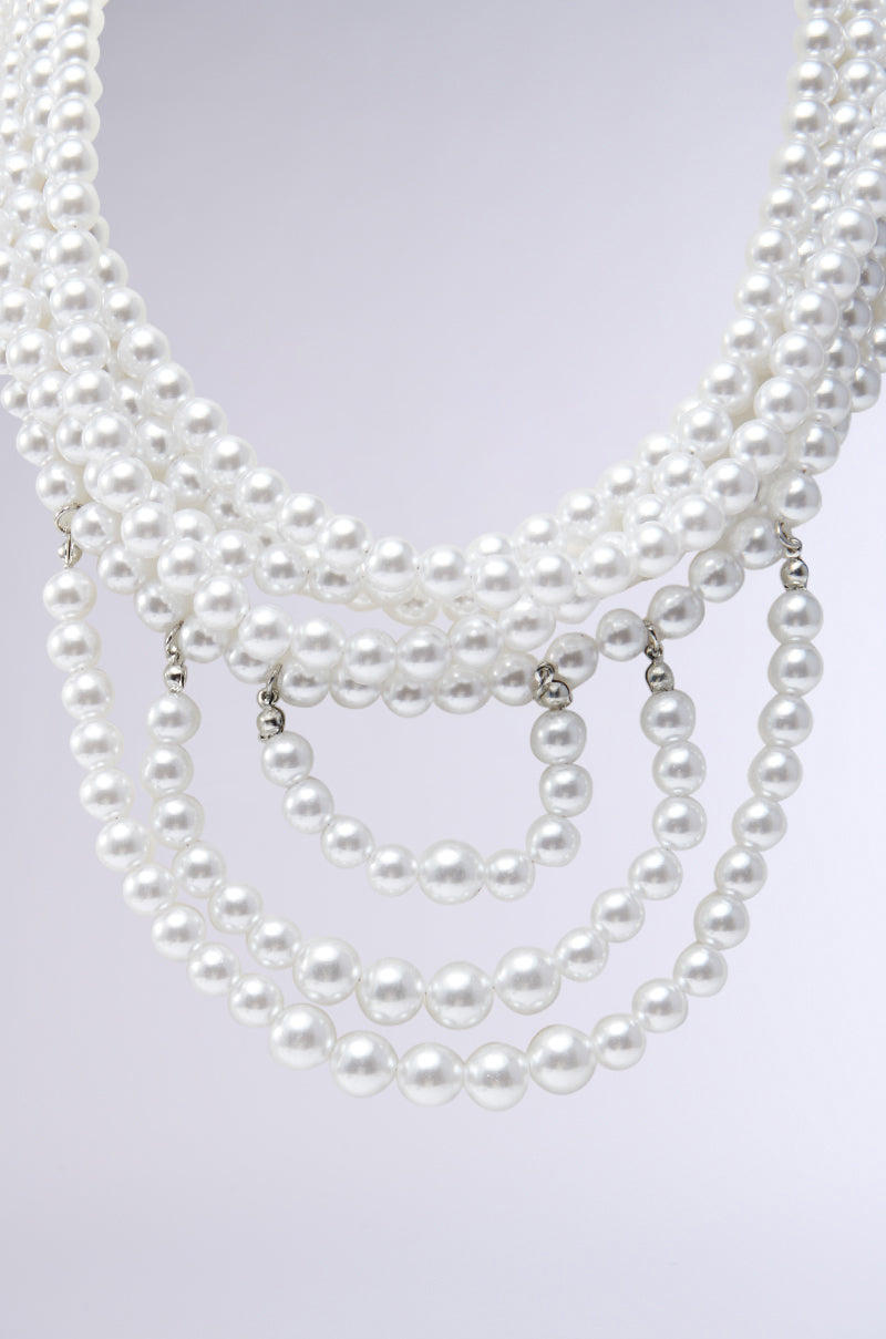 A close-up of the ANNE LAYERED PEARL NECKLACE reveals stunning multi-strand layers of white pearls, elegantly cascading in varying sizes against a neutral gray background.