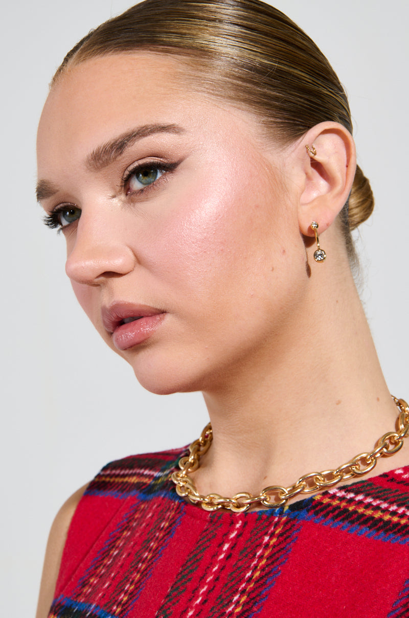 A woman with slicked-back hair and rhinestone-studded MADE YOU LOOK EARRING glances to the side. She wears a red plaid outfit, a thick gold chain necklace, complemented by subtle makeup and winged eyeliner.