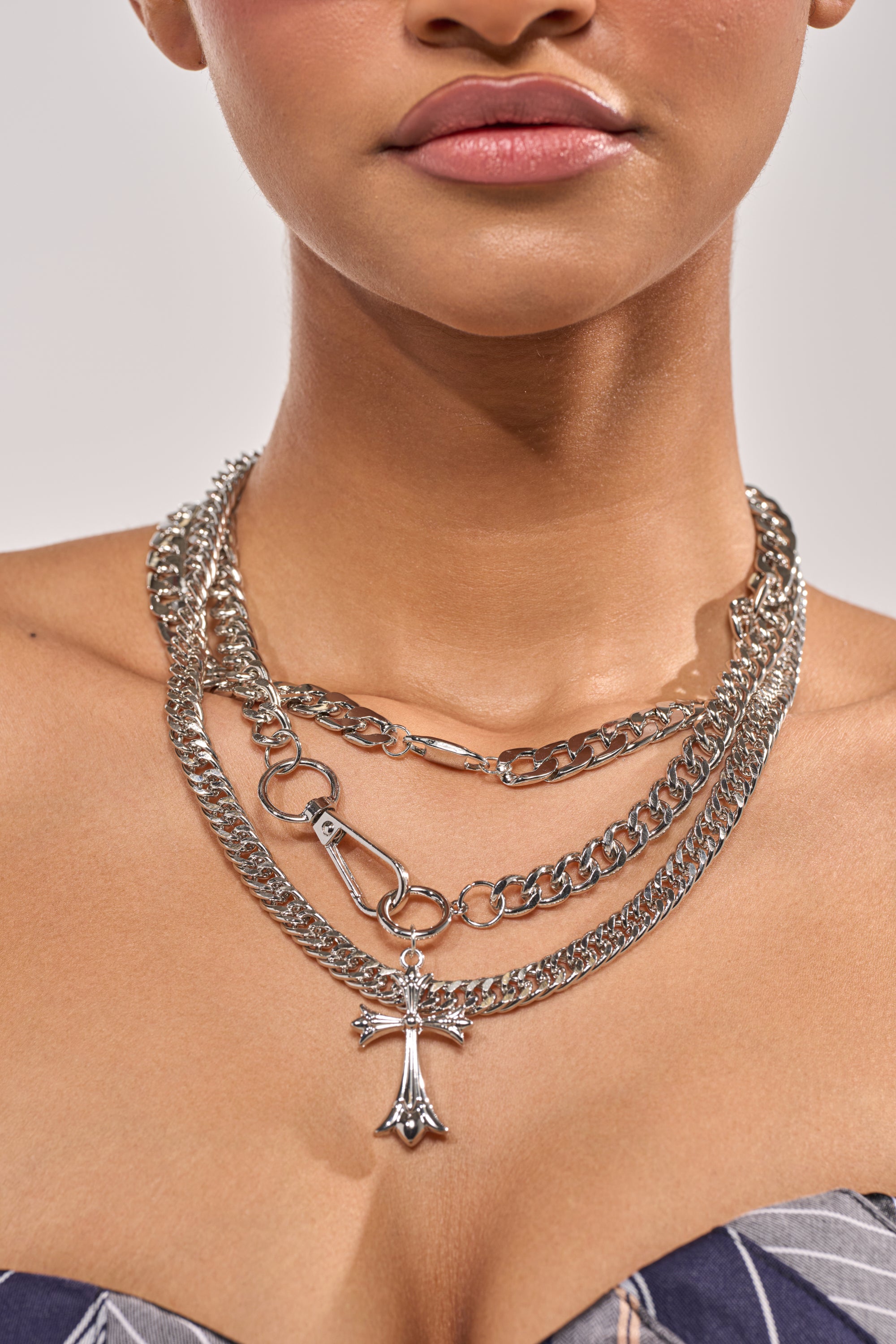 A woman wears the BLESSED TO BE HOT SILVER NECKLACE—a layered chain with a cross pendant—her bare upper chest and shoulders visible against a plain background. 1