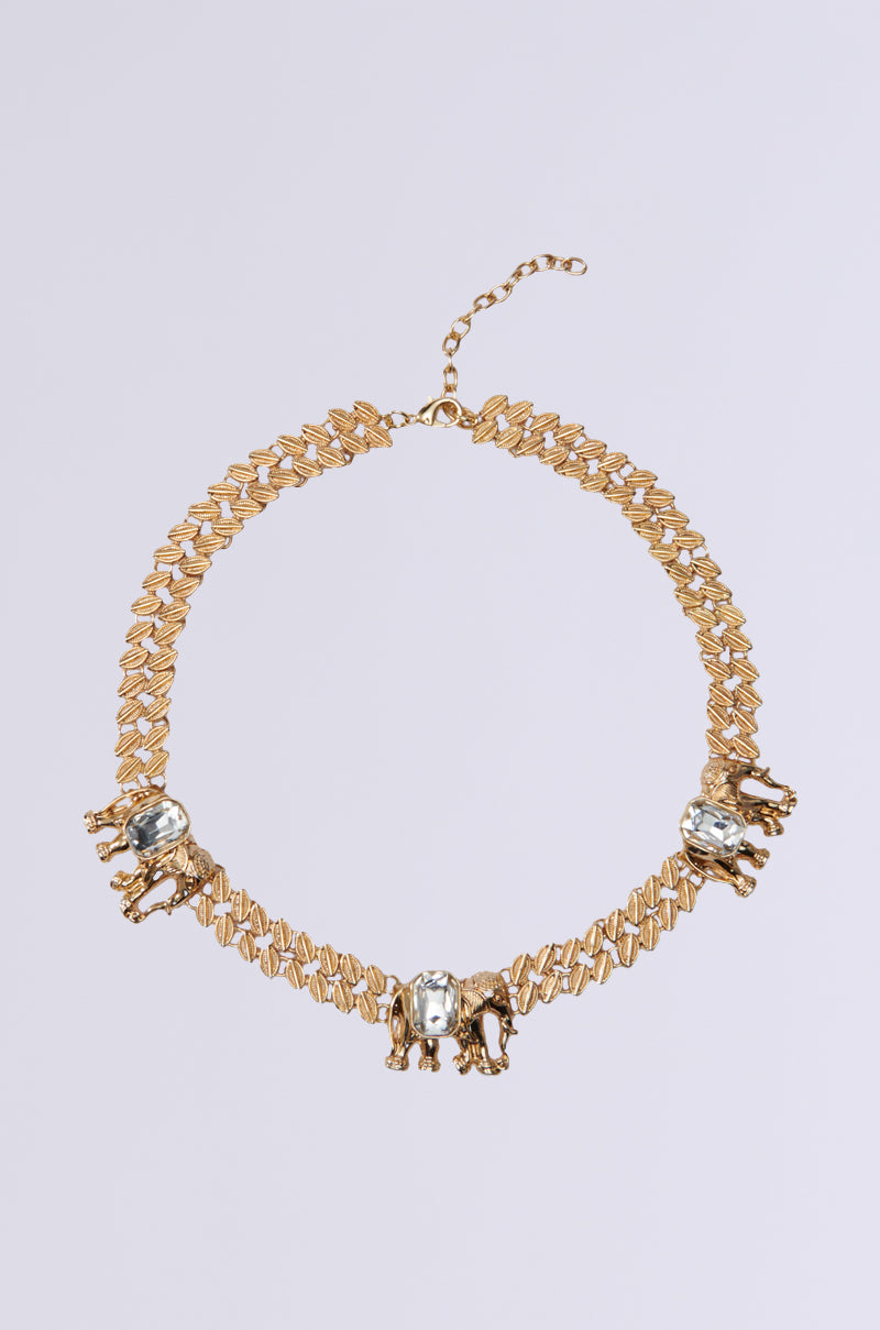 The JOANN NECKLACE is a faux gold metallic piece with a chain link design, featuring three small elephant pendants. Each pendant has intricate gemstone centers, adding elegance. The necklace includes an adjustable clasp and is displayed against a plain light gray background.