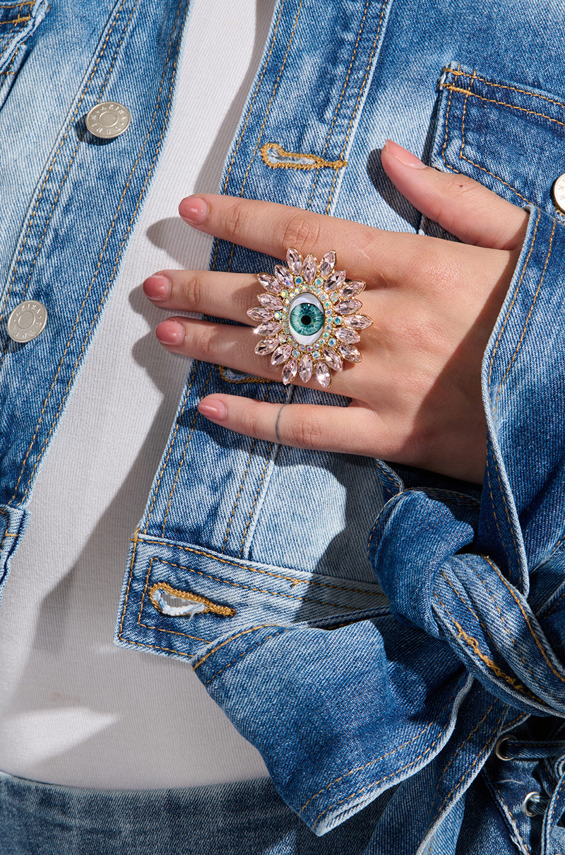 Sporting a denim jacket and white shirt, an individual showcases the "EYE SEE YOU STATEMENT RING," a striking piece featuring an eye accent design adorned with sparkling gemstones.