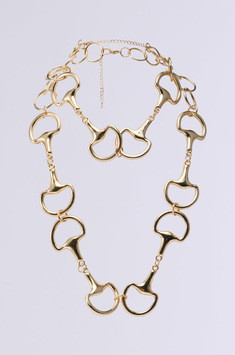The BIT MUCH NECKLACE showcases a gold chain adorned with large, abstract linked shapes against a minimalist background. The links exhibit wavy, curved forms that contribute to its artistic and modern appeal. It includes adjustable lobster clasps to ensure the perfect fit.