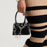 A person wearing the VIBES PANT AND PURSE CHAIN ensemble features a black and beige striped skirt while holding a small purse adorned with a metallic faux silver chain. They accessorize with a silver bracelet on their wrist and complete the look with tall black boots, creating an effortlessly chic appearance.