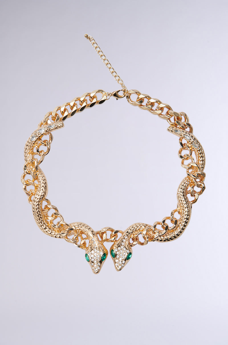 The WATCH OUT FOR THE SNAKES NECKLACE showcases two intertwined gold snake heads with faux emerald eyes and rhinestones as the front closure, while the chain links exhibit intricate detailing against a plain, light gray backdrop.