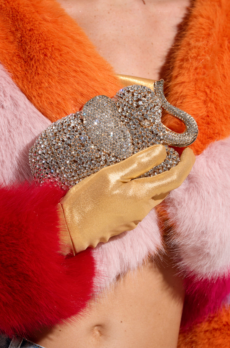 A person wearing a colorful faux fur coat and a gold glove holds an AKIRA Label BLING ELEPHANT CLUTCH. The vibrant outfit features orange, pink, and red sections, with the midriff slightly visible.