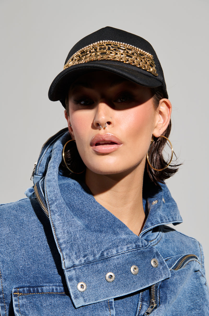 A person wearing a denim jacket and the THERES LEVELS CHAIN HAT, adorned with rhinestone detailing, looks forward. They have hoop earrings and a nose piercing. The image features a neutral gray background.