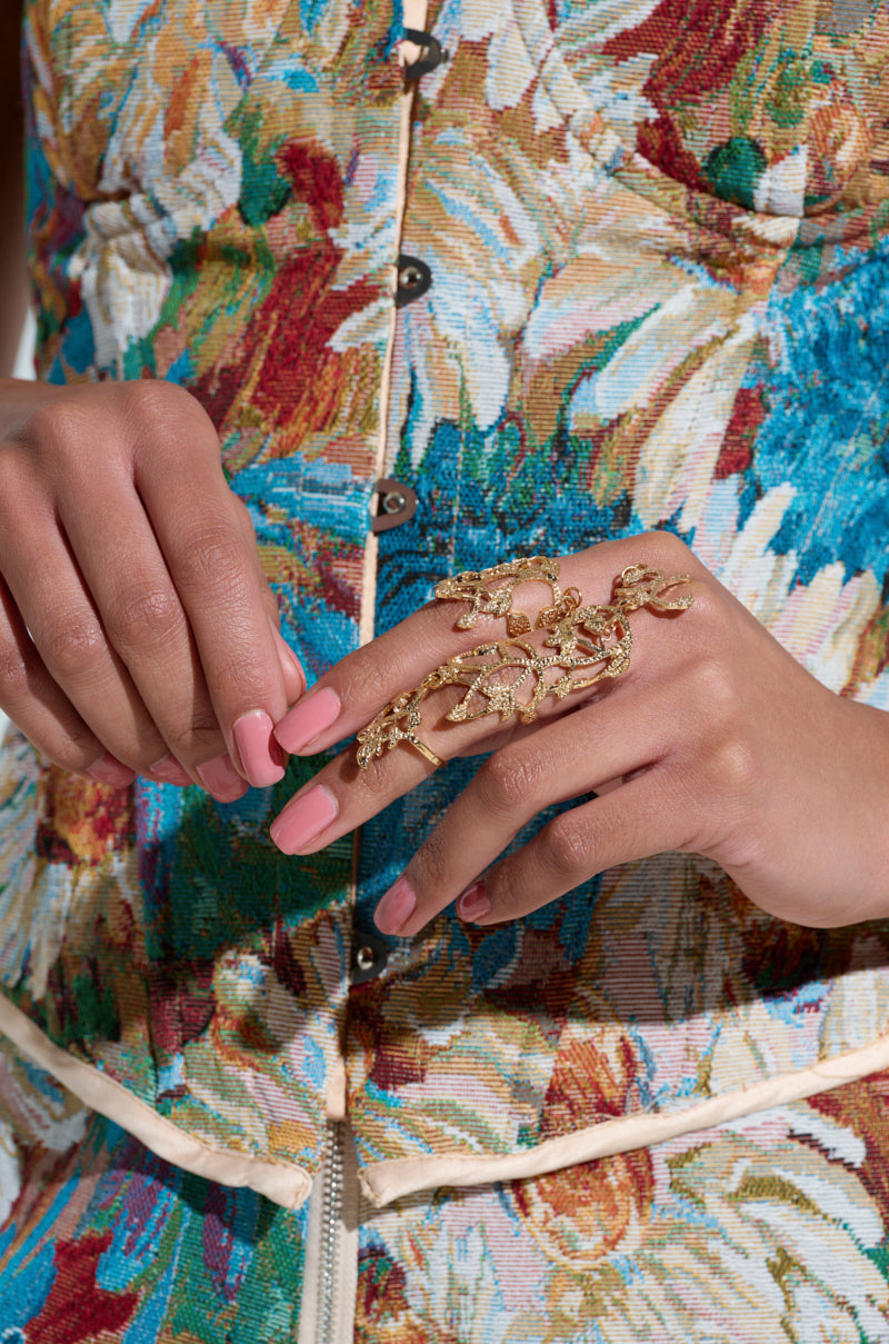 Someone flaunts an AKIRA Label ensemble, vibrant with a floral design, adorned with pink nail polish and a striking EVA TWO FINGER RING SET in ornate gold, perfectly elevating their colorful button-down outfit.