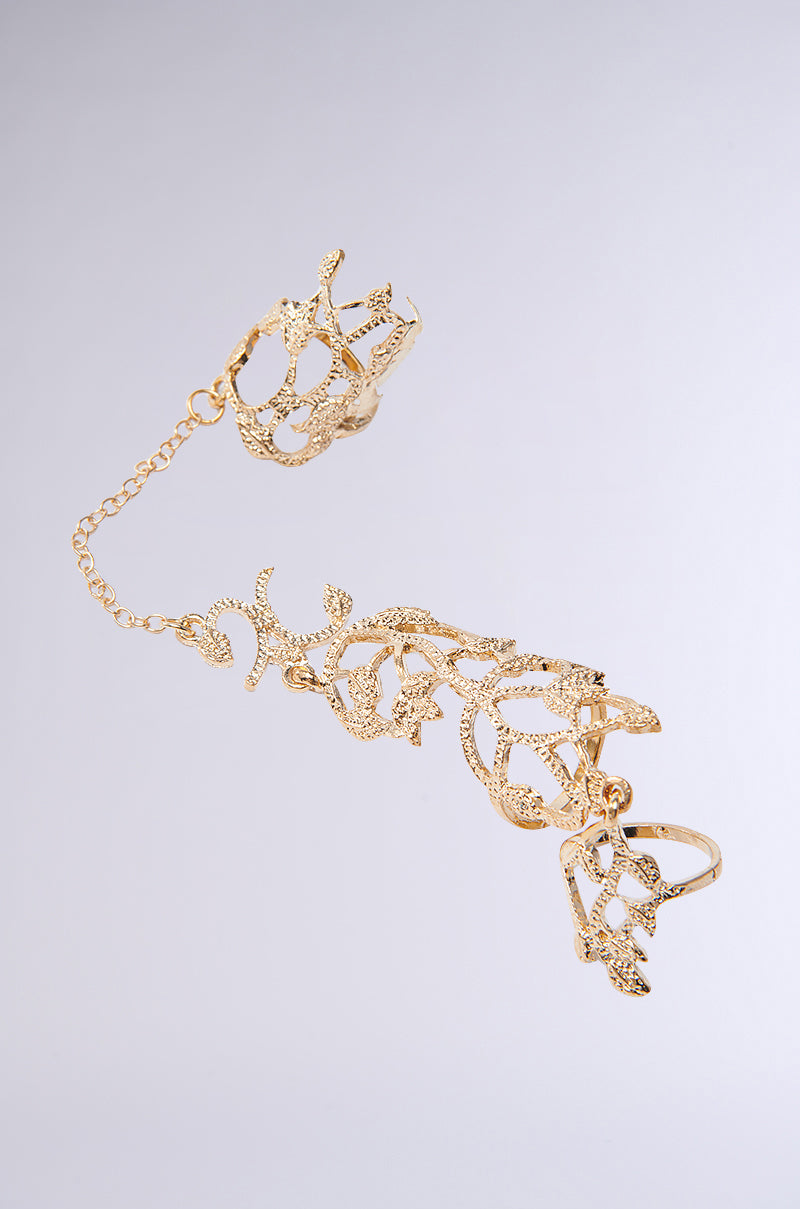 The EVA TWO FINGER RING SET features an exquisite gold hand bracelet with a delicate chain connecting to a ring, showcasing an elegant floral design and ornate lace-like patterns. It's presented against a sleek white-to-light-gray gradient background.