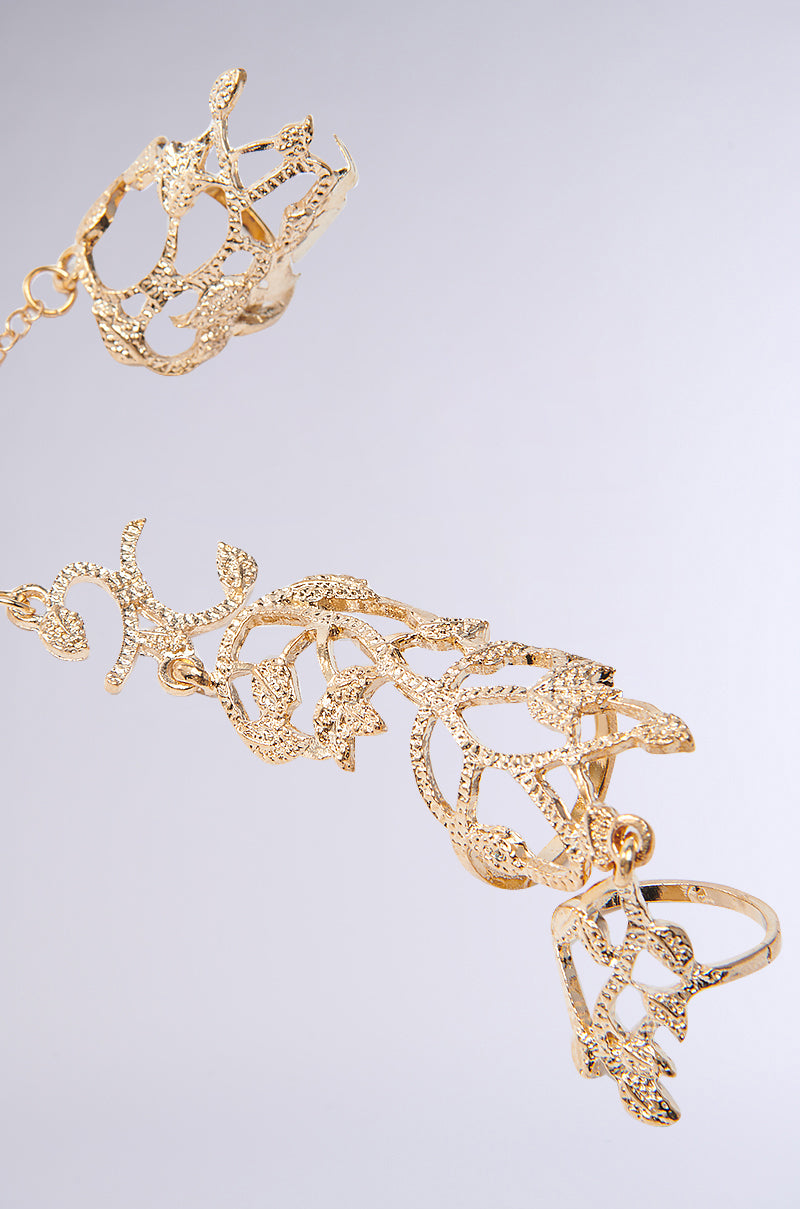 The EVA TWO FINGER RING SET showcases gold filigree hand jewelry with intricate lace-like patterns. Rings are linked by delicate chains in an ornate design featuring a subtle floral motif, set against a light gray background, embodying the AKIRA Label's distinguished style.