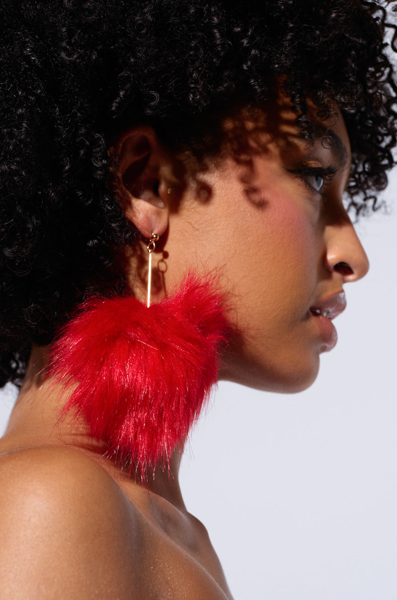 A person with curly hair is shown in a close-up profile view, wearing a large, fluffy red MAMI FUR EARRING that features classic post back closures. The background is plain, highlighting the person's side profile and the vibrant earring. 0