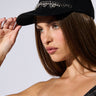 A woman with long brown hair is wearing a SPIKED SNAPBACK adorned with rhinestone embellishments and a black off-the-shoulder pinstripe top. She holds the brim of the cap with her right hand and gazes thoughtfully into the distance against a plain light background.