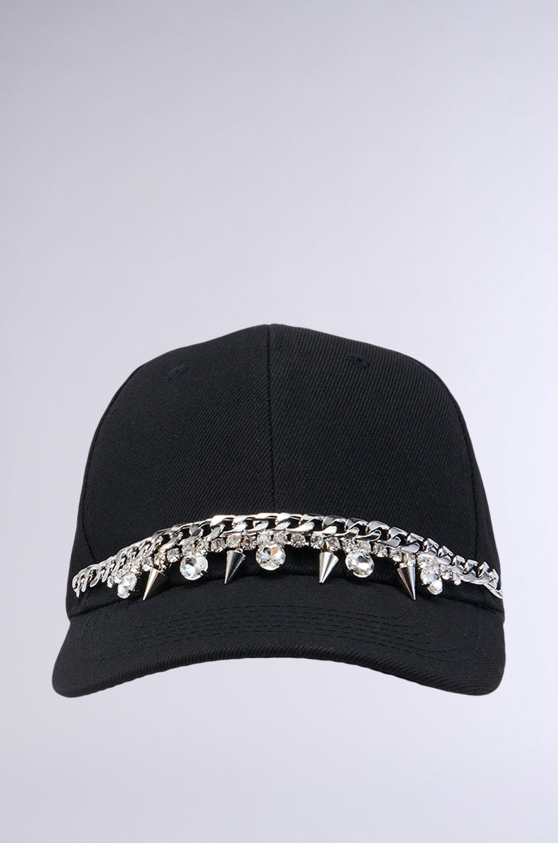 The SPIKED SNAPBACK is a black cap adorned with a striking chain of silver spikes and clear rhinestones across the front. This simple yet edgy design is complemented by an adjustable Velcro strap for the perfect fit, all set against a plain, light gray background. 2