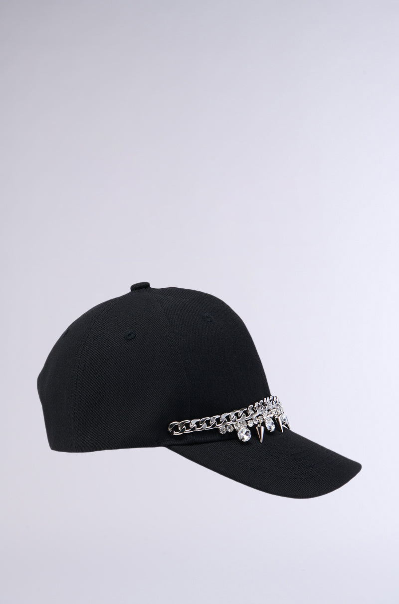 The SPIKED SNAPBACK is a black baseball cap featuring a curved brim and an adjustable Velcro strap for the perfect fit. Its simple design is starkly contrasted by edgy silver chain and spike embellishments across the front, making it reminiscent of classic spiked snapbacks. Photographed against a plain, light-colored background, this cap stands out as a bold fashion statement. 3