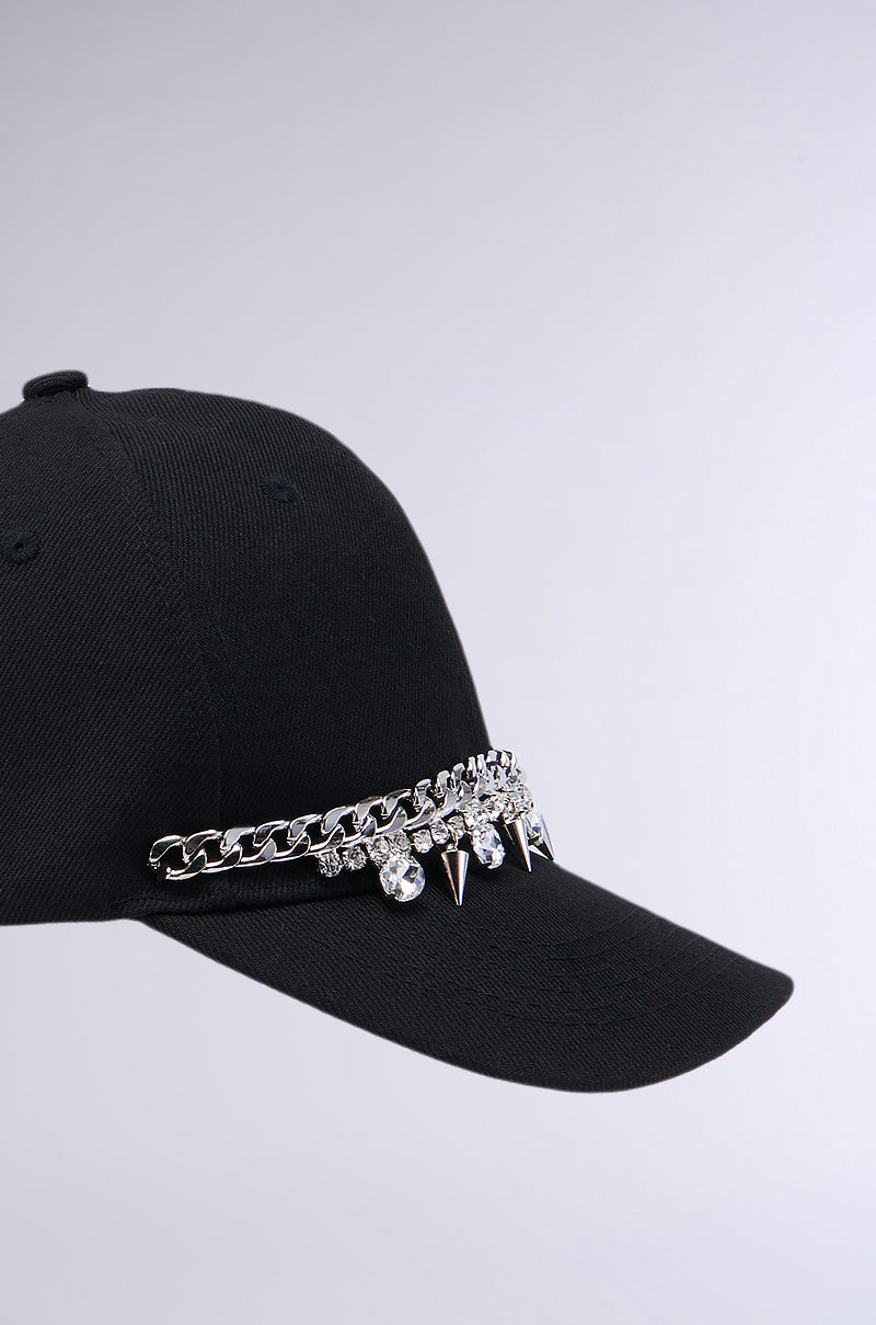 The SPIKED SNAPBACK is a black cap accented with a silver chain, spikes, and rhinestone embellishments wrapping around the front. Its simple and sleek design contrasts sharply with its edgy, eye-catching accessories and includes an adjustable velcro strap for a perfect fit. 4