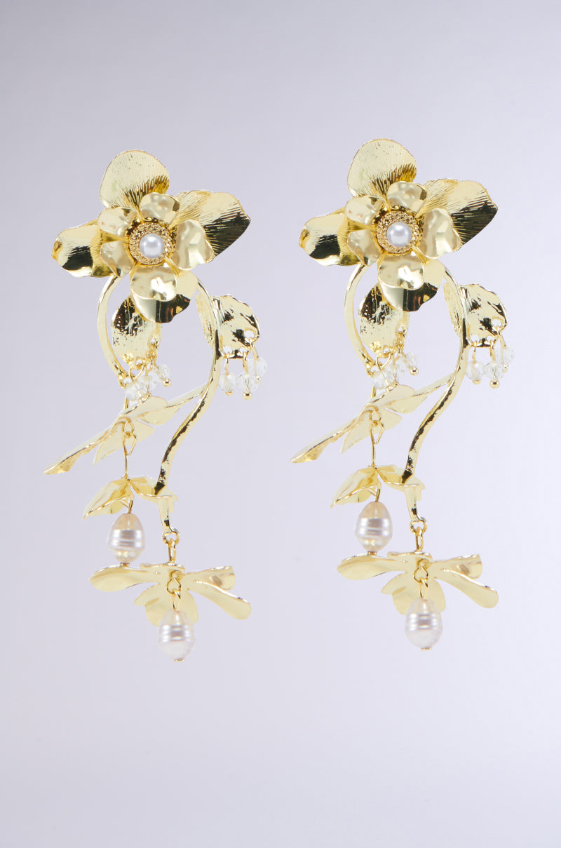 The BLOOM WITH ME DANGLE EARRING features a pair of gold floral earrings with intricate petal designs, adorned with small pearls in the center. Delicate gold stems hang below, ending with dangling pearl buds and flower accents, creating an elegant and luxurious appearance against a plain, light gray background.