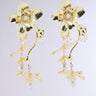 The BLOOM WITH ME DANGLE EARRING features a pair of gold floral earrings with intricate petal designs, adorned with small pearls in the center. Delicate gold stems hang below, ending with dangling pearl buds and flower accents, creating an elegant and luxurious appearance against a plain, light gray background.